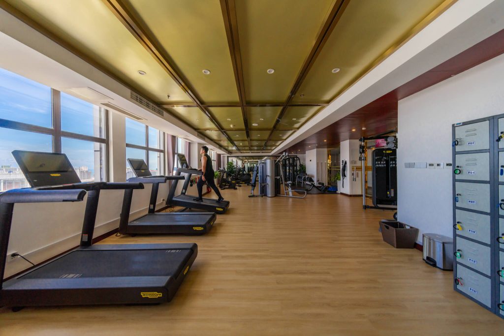 Full view of the gym at Legend Hotel & Resort Sihanoukville, Cambodia. Treadmills, weight lifting equipment are visible, along with a locker room area.